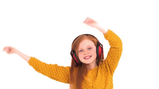 Close-up of a little cheerful girl with charming smile and magical foxy hair listening to the music in wireless headphones and dancing. Happy emotions concept. Isolated, on white background
