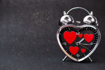 Steel clock alarm in the form of a heart with red hearts inside on a black glitter shiny background. Happy Valentine's Day
