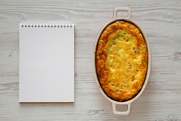 Homemade Cheddar Corn Pudding Casserole, blank notepad on a white wooden background, top view. Flat lay, top view, from above. Space for text.