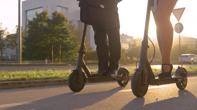 LENS FLARE, LOW ANGLE, CLOSE UP: Detailed shot of e-scooters as two business people commute home on a sunny evening. Work colleagues ride eco friendly electric scooters to work at scenic sunrise.