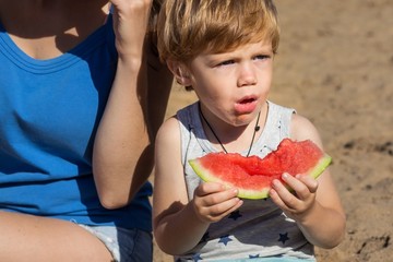 little redhead boy chewing juicy piece of watermelon sitting on the sand near his mother and looking with surprise in summer sunny day