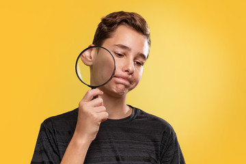 The concept of teenage acne. A teenage boy with a disgruntled face, holding a magnifying glass near his cheek. Yellow background. Copy space