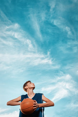 Fototapeta premium Sports and basketball. A young teen in a black tracksuit playing basketball on the school Playground. Cloudy blue sky on the background. View from the bottom. Vertical. Copy space