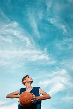 Sports And Basketball. A Young Teen In A Black Tracksuit Playing Basketball On The School Playground. Cloudy Blue Sky On The Background. View From The Bottom. Vertical. Copy Space