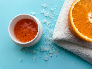 A jar of orange scrub, a half of orange on a folded white towel and scattered around a large bath salt on a soft blue background. Spa concept, body cleansing and moisturizing, anti-cellulite
