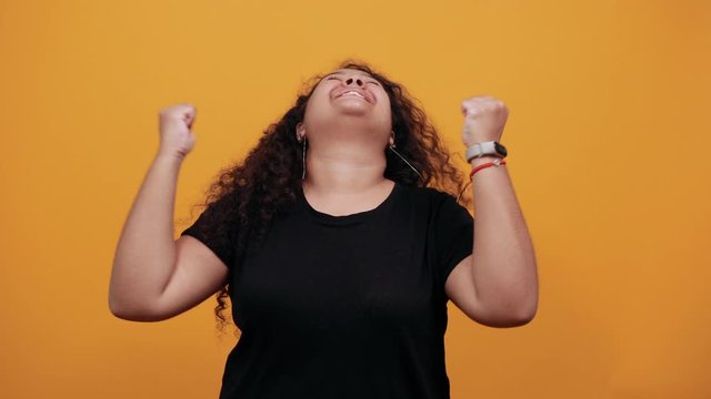 Happy afro-american young woman with overweight keeping fists up, smiling, dancing over isolated orange background wearing fashion black shirt. People lifestyle concept.