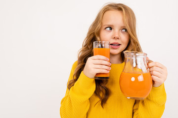 Happy teenager girl with red hair, hoody and yellow trousers drinks carrot juice isolated on white background