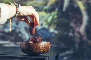 Woman hand with royal dates fruit in a bowl of coconut on a table in a palm tree grove.