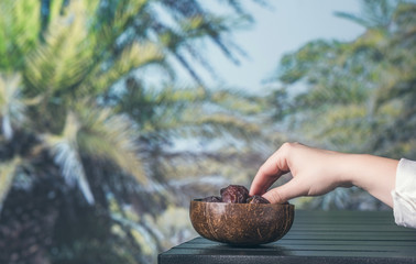 Woman hand with royal dates fruit in a bowl of coconut on a table in a palm tree grove.