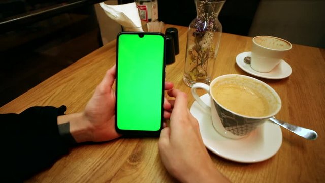 Young female hands using green screen smartphone sitting in cafe drinking coffee. Online business communication. Chromakey mock-up. Tapping empty touchscreen. Social network. Internet chatting. 