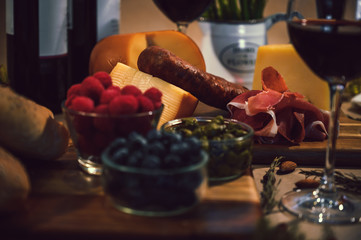 Table with gourmet ingredients, glass of wine, variety of cheeses, cold meats, grapes, raspberry, blackberry and artisan bread on a rustic background