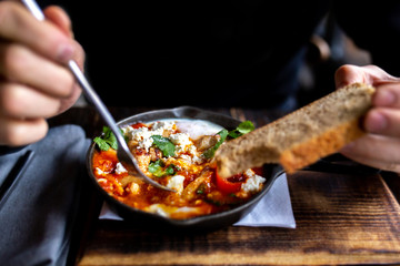 Shakshuka with eggs, sausages, cheese and parsley in a pan. Shakshuka - traditional Israeli tomato stew with egg. A small frying pan with food on a wooden board. A man eats from a carpet with a fork.