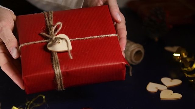 Close up on a women wrapping gifts for Valentine's Day in red craft paper. The gift is decorated with strings and heart-shaped wood decorations