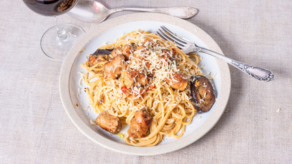 Spaghetti with homemade sausages and eggplant sprinkled with grated cheese in a plate and a glass of red wine on the table - a traditional Italian rural lunch