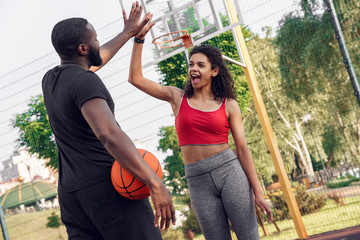 Outdoors Activity. African couple standing on basketball court man back view with ball giving high...
