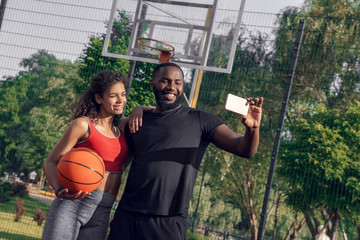 Outdoors Activity. African couple standing on basketball court boyfriend taking selfie on smartphone with girlfriend smiling happy