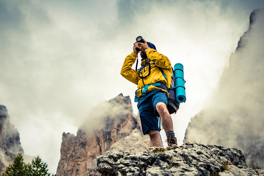 Young man hiker taking pictures on stone mountain with cloudy sky and fog. Yellow jacket, backpack, black beard and beanie. Traveling Dolomites, Italy. - Powered by Adobe