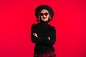 Portrait of young pretty woman on red background in studio. Trendy hipster girl in black hat, sunglasses and plaid mini skirt. Smiling lady.