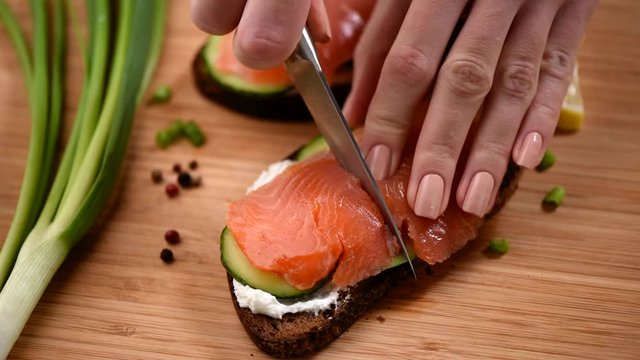 Cut fish sandwich in two halves. Woman cutting rye bread sandwich with smoked salmon, cucumber and cream cheese in two halves. Cooking process