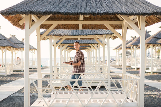 A Handsome Young Man Is An Outsourcing Employee Working With A Laptop Under A Beach Canopy On A Sunny Summer Day. The Concept Of Work And Leisure At The Same Time