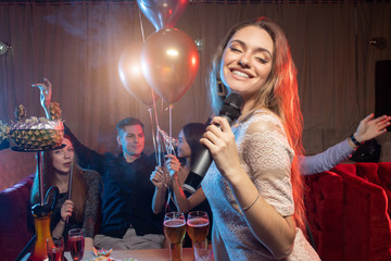charming female with blond hair emotionally sing in microphone in karaoke bar while friends...