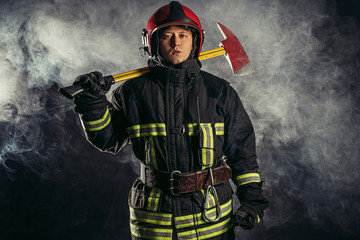 Fototapeta premium young caucasian handsome and strong fireman in uniform wearing helmet on head hold hammer in hands, going to save people from fire