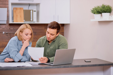young caucasian couple search bills and expenses on mortgage using laptop at home, sit together and look at screen of laptop, with documents on table