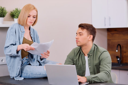 spouses sitting with modern laptop and documents, analyzing paperwork and house utility bills, young couple consider papers managing finances and expenses, checking e-mail and surfing the Net at home