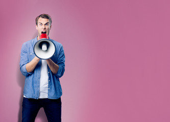 Fototapeta premium Excited man shouting through megaphone, with copy space for slogan, advertising or text, over pink background. Caucasian male model in blue casual clothing making announcement.