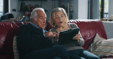 A happy mature senior couple is making a selfie or video technology call to their relatives with a tablet in living room at home. - Powered by Adobe