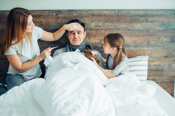 young caucasian man suffer from disease, have high temperature, lying on bed with careful kind wife and child girl. Sick man on bed