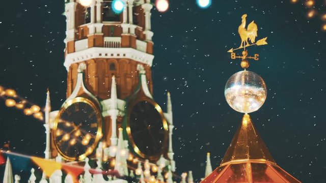 Kremlin chimes in snowy weather. Close up view to the Moscow Kremlin Clock on Spasskaya Tower through the colorful New Year garlands, shiny disco ball and wind vane of the decorated Red Square on