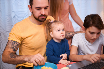 Naklejka premium adorable kids and careful parents sit in kitchen with laptop, watching films and videos, eating fruits, having breakfast together at morning