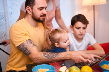 lovely adorable caucasian family consisted of young mother with long hair, bearded father and little kids son and daughter sit together on table, using laptop and eating, having meal, breakfast