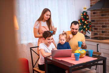 adorable kids and careful parents sit in kitchen with laptop, watching films and videos, eating fruits, having breakfast together at morning