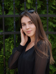 Fototapeta premium Outdoor summer lifestyle portrait of young beautiful brunette woman in black dress enjoying warm weather posing near fence city park with adorable smiling touching face
