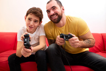 Fototapeta premium young caucasian man with beard sit with son on sofa and play video game together, holding joysticks, wearing casual t-shirt. indoors