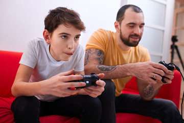 joyful playful father and son playing video game sitting on red sofa, happy parent and kid boy enjoy spending time together