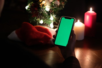 over shoulder of one hand holding green screen smartphone. Blur christmas tree, hat and candle lights. black background