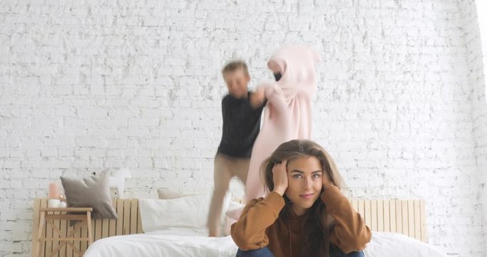 Hyperactive preschool boy son with attention deficit hyperactivity disorder ADHD exasperates exhausted mother jumping on bed, mom cover ears with hands suffers lack of energy, tired of noise.