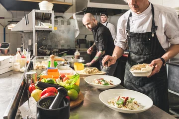 Foto op Plexiglas Professionele chef-kok die in het keukenrestaurant van het hotel kookt en het diner voorbereidt. Een kok in een schort maakt een salade van groenten en pizza. © xartproduction