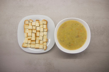 square croutons on a white plate next to a round white bowl of soup on a gray kitchen table with texture top view