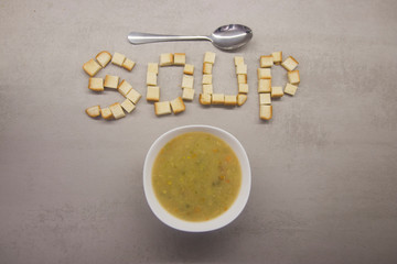 word SOUP made of square toasts next to a round bowl of soup and a spoon on a gray kitchen table with texture top view