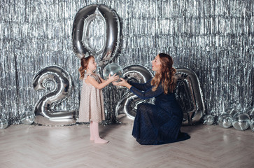 Obraz na płótnie Canvas Stock photo of beautiful mother in dark blue dress giving a silver shining disco ball to her lovely daughter in diadem and festive beige dress. They are standing against inflated numbers of New Year