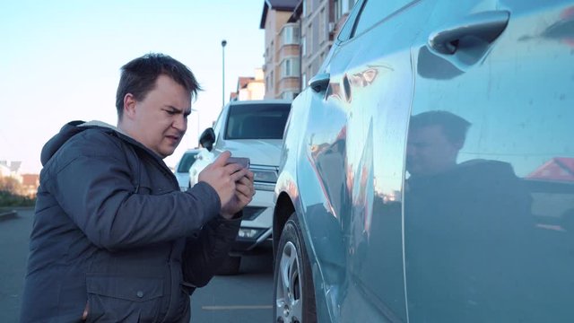 Man is sitting with phone on ground in parking lot on street and taking photos of dent on car door for insurance company. Guy is upset because of accident, sadly stroking door with hand, then leaving.