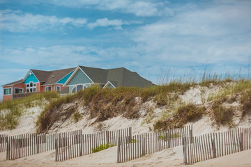 Big bright colorful ocean side beach house Outer Banks, North Carolina. Sand dunes, beach grass and wooden fence in the foreground, and clear blue sky with clouds in the background.