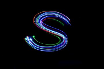 Long exposure photograph of a letter s in neon colour in an abstract swirl, parallel lines pattern against a black background. Light painting photography.