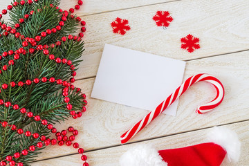 Empty greeteing card and sweet candy cane on a wooden table near a fir branch with red bead garland and Santa hat. Christmas and New Year holydays.