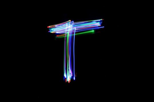 Long exposure photograph of a letter t in neon colour in an abstract swirl, parallel lines pattern against a black background. Light painting photography.