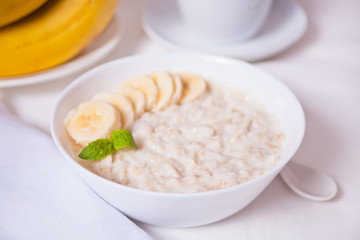 Oatmeal porridge with banana in the white bowl on the white table. Health breakfast.